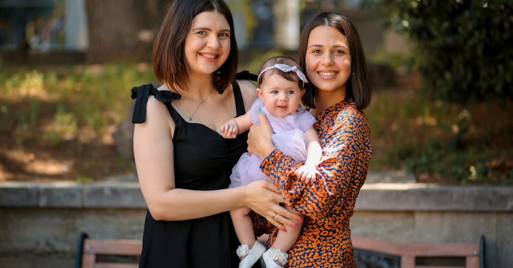 Two women holding a baby, smiling warmly in a sunny park setting.